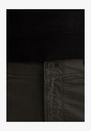 Detailná záber tmavozelených nohavíc so zapínaním na kovový gombík s logom pod čiernym rebrovaným pásom.