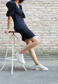 Svart figurnära klänning med puffärmar, matchad med vita sneakers med ett blått mönster. Bakgrund: texturerad tegelvägg.