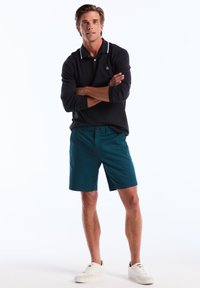 Polo à manches longues bleu marine avec des rayures blanches au col ; short turquoise ; le modèle porte des baskets blanches ; debout les bras croisés, fond clair.