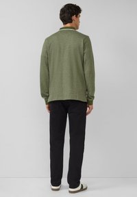 Zielona, teksturowana koszulka polo z długim rękawem i kontrastowym kołnierzem, w zestawieniu z czarnymi spodniami i casualowymi butami, widziana od tyłu.