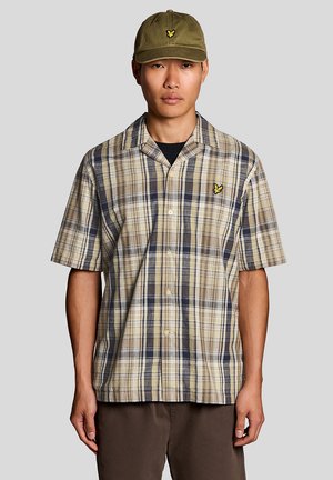 Homme portant une casquette vert olive et une chemise à manches courtes à carreaux beige, marine et marron avec un logo d'oiseau jaune sur la poitrine.