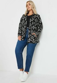 Giacca a scacchi bianco e nero con design geometrici, dotata di zip frontale e tasche laterali, abbinata a jeans blu e sneaker bianche.
