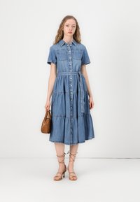Robe en denim à manches courtes, boutonnière devant, jupe à volants et taille ceinturée. Couleur bleu clair, avec deux poches poitrine.