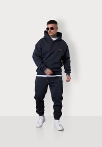 Sweat à capuche bleu marine avec poche avant et logo brodé, associé à un jogging assorti. Il présente des poignets élastiques et un cordon de serrage sur le pantalon.