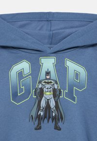 Μπλε φούτερ με κουκούλα που έχει εικόνα υπερήρωα με γκρι και μαύρη στολή. Η λέξη «GAP» τυπωμένη με έντονα πράσινα γράμματα πάνω από την εικόνα.