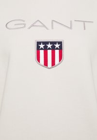 Bílá mikina s vyšitým logem GANT v šedé barvě a štítkem s červenými a modrými pruhy a třemi bílými hvězdami.