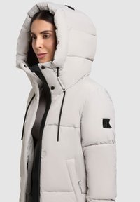 Light gray padded jacket with a large hood, black zippers, and elastic drawstrings. Features a side pocket and a logo on the sleeve.