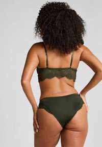 Vrouw met krullend haar, draagt een donkergroene beha met kant en bijpassend ondergoed, staat met één hand op de heup tegen een effen achtergrond.