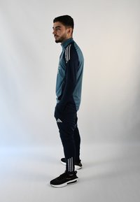 Conjunto deportivo que incluye una camiseta de manga larga en color verde azulado con rayas blancas en los hombros, combinado con pantalones azul marino y zapatillas negras.