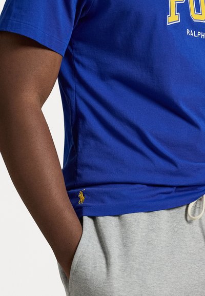 Blue cotton t-shirt with ribbed texture, featuring white "Polo" branding and a small yellow embroidered logo at the hem, paired with gray sweatpants.
