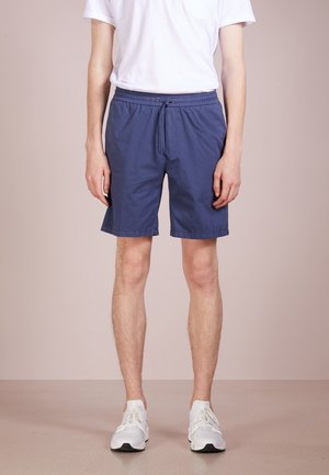 Homme portant un short bleu marine, un t-shirt blanc et des baskets blanches, debout devant un fond beige uni.