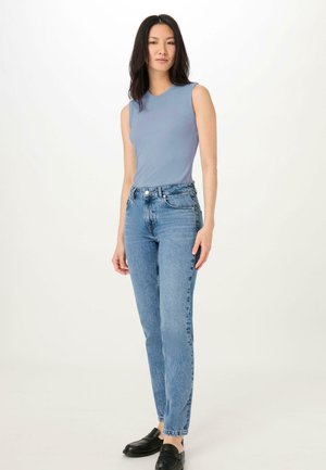 Blue sleeveless top paired with high-waisted light blue jeans. Model wearing black loafers, standing on a neutral background.