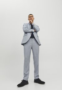 Lichtblauw pak met een slim fit, voorzien van een enkelvoudig gebreide blazer, platte broek en zwarte schoenen, tegen een neutrale achtergrond.