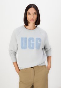 Light grey sweatshirt with a plush blue "UGG" logo, round neckline, and ribbed cuffs, paired with light brown tailored pants.