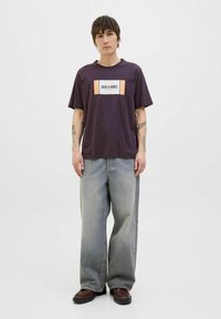 Camiseta corta de manga corta púrpura con logo gráfico en blanco y naranja, combinada con jeans de pierna ancha de mezclilla azul deslavada y zapatos marrones. Fondo neutro.