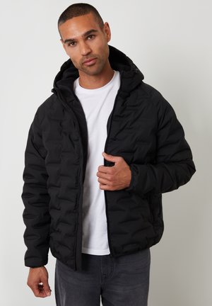 Schwarze gepolsterte Jacke mit Reißverschluss und Kapuze. Verfügt über eine rautenförmige Stepptextur und elastische Bündchen. Getragen über einem weißen T-Shirt.