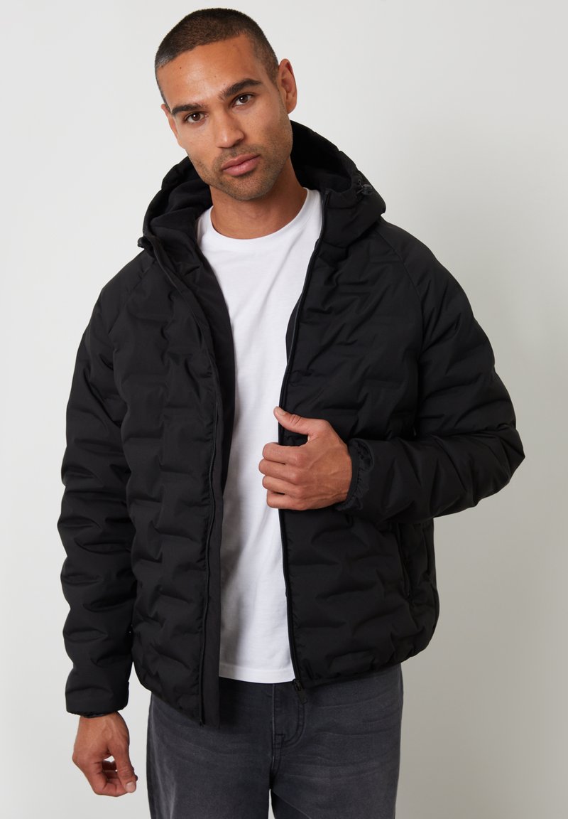 Giacca nera imbottita con zip frontale e cappuccio. Presenta una texture a rombi e polsini elasticizzati. Indossata sopra una t-shirt bianca.