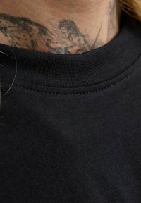 Kaklo tatuiruotė su mažu tamsiu figūromis, turinčiais garbanotus plaukus ir ištiesinta ranka, dalinai matoma virš juodo marškinių krašto.