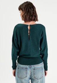 Pull en tricot vert avec une ouverture en forme de clé au dos, fermeture à bouton, ourlet côtelé et manches longues, associé à un jean bleu délavé.