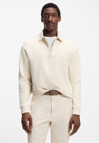 Beiger Sweatshirt mit Viertel-Reißverschluss am Kragen und Brusttasche, kombiniert mit passenden Hosen und einem braunen Gürtel. Glatte Textur.