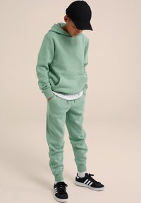Felpa in cotone verde menta con tasca frontale e cappuccio con coulisse, abbinata a pantaloni della tuta coordinati e sneakers nere con strisce bianche.
