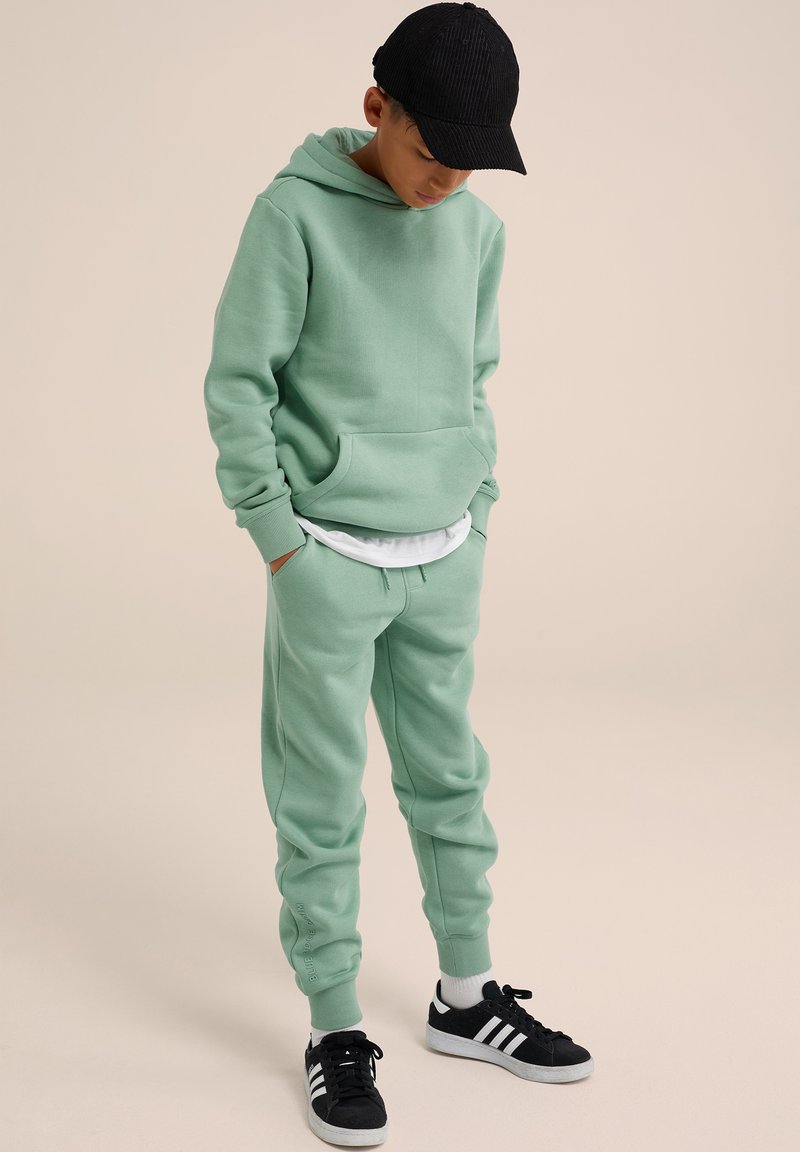 Felpa in cotone verde menta con tasca frontale e cappuccio con coulisse, abbinata a pantaloni della tuta coordinati e sneakers nere con strisce bianche.