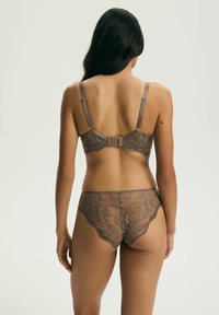 Žena s dlhými tmavými vlasmi, ktorá má na sebe hnedú kvetinovú čipkovú podprsenku a k nej ladúce čipkované nohavičky, zobrazená z chrbta na jednoduchom pozadí.