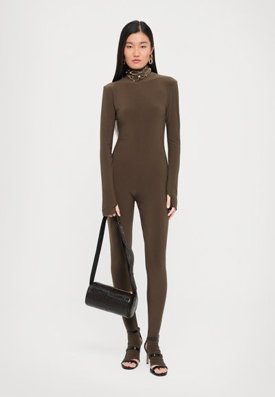 Brown long-sleeve jumpsuit with a high collar, soft texture, and slim fit. Accessorized with a black handbag and open-toe heels.