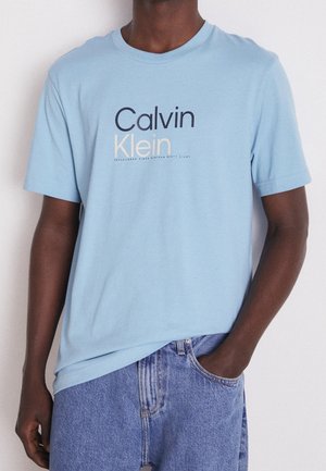 Mężczyzna ubrany w jasnoniebieską koszulkę Calvin Klein i niebieskie jeansy, z jedną ręką w kieszeni, na jednolitym tle.