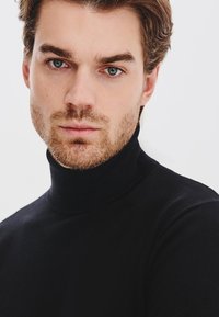 Czarny sweter z golfem o gładkiej fakturze, dopasowany fason i wysoki kołnierz. Model ma jasnobrązowe włosy i zarost. Neutralne tło.