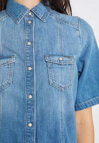 Camisa de mezclilla en azul claro, con cuello de punta, mangas cortas y dos bolsillos en el pecho con cierres a presión. Detalles de costura notables.