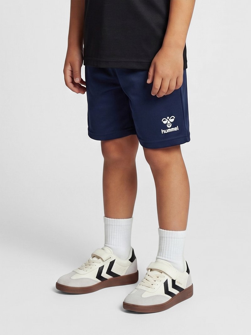 Kind draagt marineblauwe shorts met een wit hummel-logo, witte sokken en witte sneakers met zwarte chevronstrepen, staand op een grijze vloer.