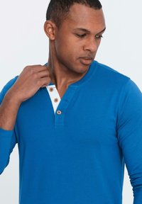 Maglietta henley azzurra a maniche lunghe con un collettino bianco e tre bottoni in legno, con una texture liscia e un design casual.