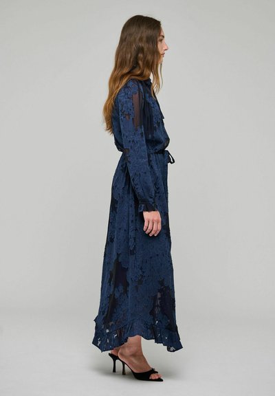 Femme vue de profil portant une longue robe en dentelle florale bleu marine à manches longues et des sandales noires à talons sur un fond gris uni.