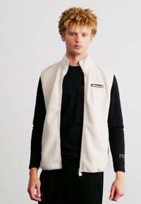 Nēšamais vestes mētelis krēmkrāsā ar rāvējslēdzēju un augstu apkakli. Valkāts virs melna garām piedurknēm krekla, uz kura krūšu daļā ir logo.