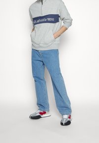 Hombre con sudadera gris de Quiksilver, jeans azul claro y zapatillas gris, rojo y blanco, de pie con las manos en los bolsillos de la sudadera.