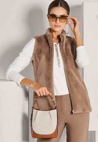 Frau mit braunem Fellwesten, weißem Langarmoberteil, braunen Hosen, großen Creolen, Sonnenbrille und einer beigen und braunen Handtasche.