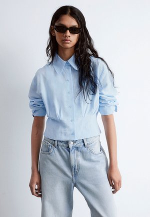 Light blue button-up shirt with a pointed collar and rolled sleeves, paired with light denim jeans. Simple design and relaxed fit.