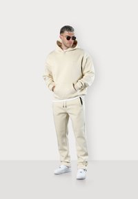 Beige hoodie with a large front pocket and drawstring, paired with matching beige joggers featuring side pockets. Worn with white sneakers.