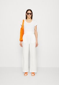T-shirt blanc à manches courtes, pantalon blanc ample, baskets oranges épaisses et un sac fourre-tout en tricot orange. Des lunettes de soleil sombres finalisent le look.