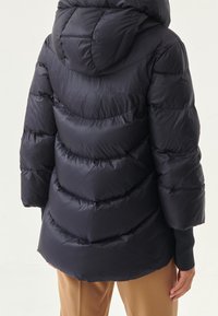 Tummanlaivansininen puffatakki hupulla, jossa on tilkkutöinä ommeltuja osia, elastiset hihansuut ja hieman ylisuuri mitoitus. Sileä, kiiltävä materiaali.