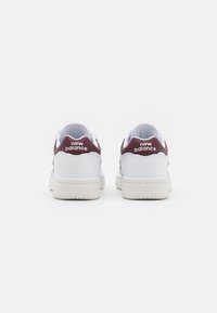 Zapatillas de piel blanca con acentos en marón en el talón, que presentan una suela de goma texturizada y el logo de "New Balance" en el talón.