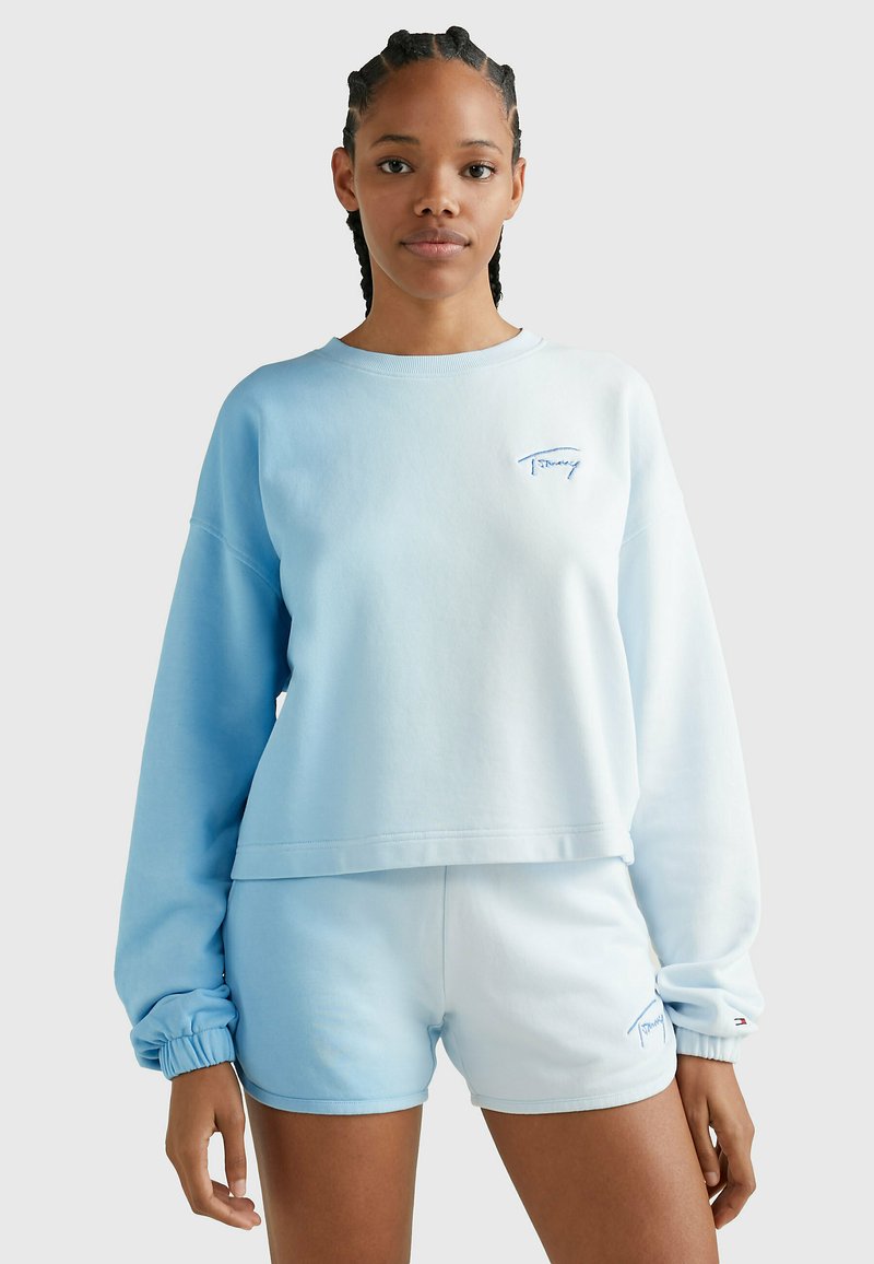 Sudadera de color azul claro con un diseño recortado y mangas largas, combinada con pantalones cortos a juego. Cuenta con un logotipo bordado y puños fruncidos.