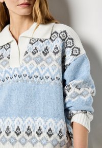 Jasnoniebieski sweter dzianinowy z białym kołnierzykiem, z geometrycznym wzorem w kolorze czarnym, szarym i niebieskim. Rękawy mankietowe oraz teksturowany materiał.