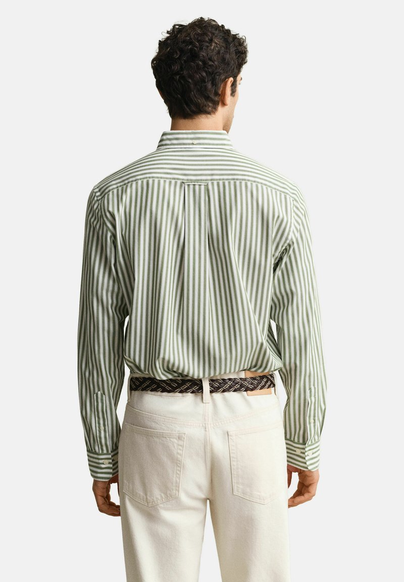 Uomo con camicia a maniche lunghe a righe verticali verdi e bianche infilata in pantaloni color avorio con cintura intrecciata marrone, visto di spalle.