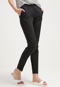 Femme portant un pantalon noir slim et une chemise rayée, debout avec les mains dans les poches et portant des sandales blanches à enfiler.