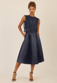 Vestito blu navy con corpetto in pizzo e bordi ondulati, caratterizzato da una vita aderente e una gonna in satin fluida. Include tasche laterali e una texture liscia.
