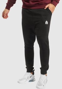 Black sweatpants with a slim fit, pockets, and a white logo on the left thigh. Made from a soft, textured fabric, detailed with ribbed cuffs.
