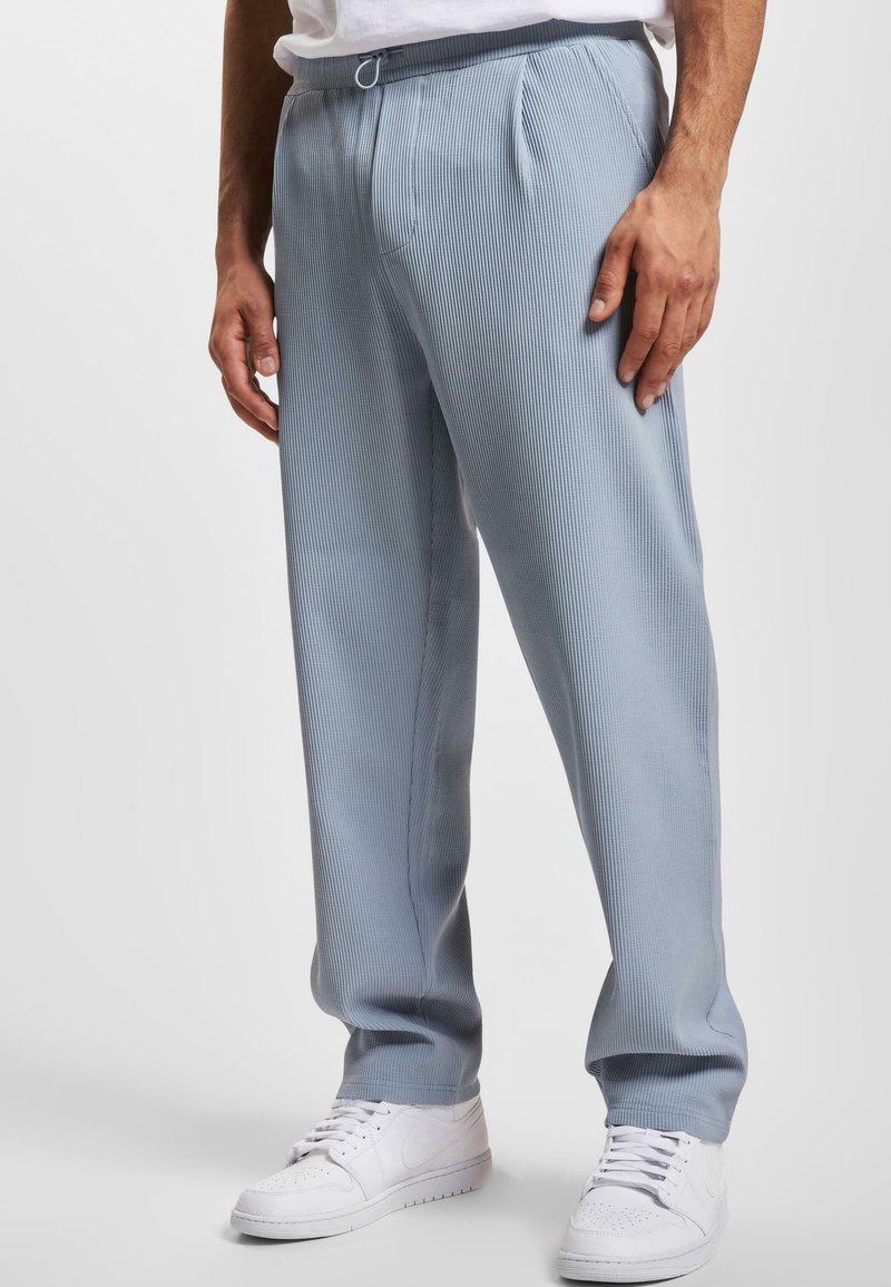 Pantalones de rayas en azul claro con cintura ajustable, que presentan pliegues frontales y un corte relajado, combinados con zapatillas blancas.