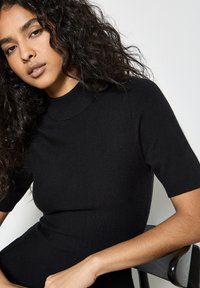 Top in maglia nera a maniche corte, aderente, con un collo alto. Texture liscia e design senza cuciture, indossato con un braccio appoggiato su una sedia.